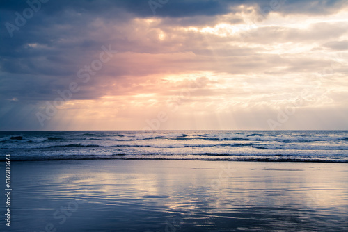 Fototapeta Naklejka Na Ścianę i Meble -  Sunset over Muriwai beach. Sun beams breaking through heavy clouds as waves splashing against the rocks. Auckland, New Zealand