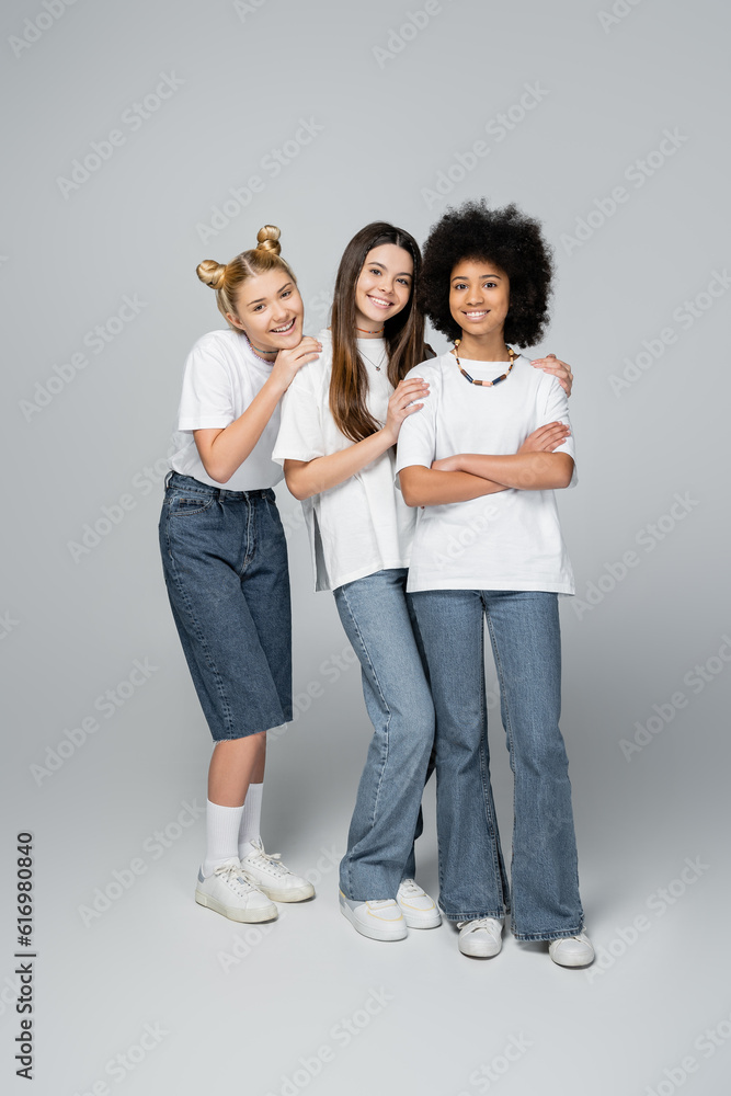 Full length of positive and interracial teenagers in casual white t-shirts and jeans looking at camera and standing on grey background, lively teenage girls concept, friendship and bonding Full length of positive and interracial teenagers in casual white t-shirts and jeans looking at camera and standing on grey background, lively teenage girls concept, friendship and bonding