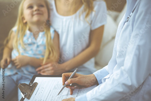 Doctor and patient. Pediatrician using clipboard while examining little girl with her mother in clinic at home. Happy cute caucasian child at medical exam. Medicine concept
