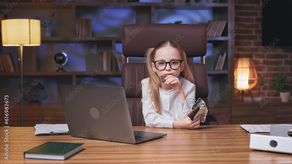 Cute little blonde girl with ponytails dressed as business lady sitting at wooden desk with remote computer and eating milk chocolate. Charming schoolkid enjoying sweets during short break in office. Cute little blonde girl with ponytails dressed as business lady sitting at wooden desk with remote computer and eating milk chocolate. Charming schoolkid enjoying sweets during short break in office.