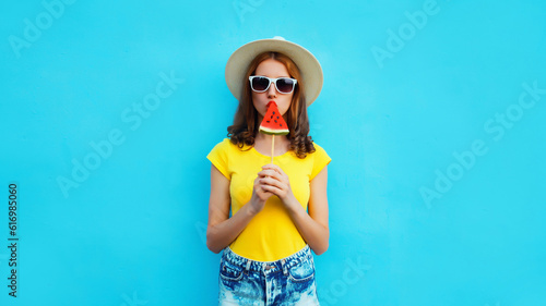 Summer portrait of young woman with sweet juicy lollipop or ice cream shaped slice of watermelon wearing straw hat on blue background