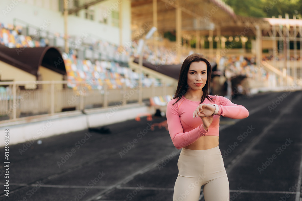 Fototapeta premium Female runner looks at smartwatch while going for run checks time listens music via headphones dressed in jacket strolls outdoor against modern tall buildings tracks wearable device wanders in city.