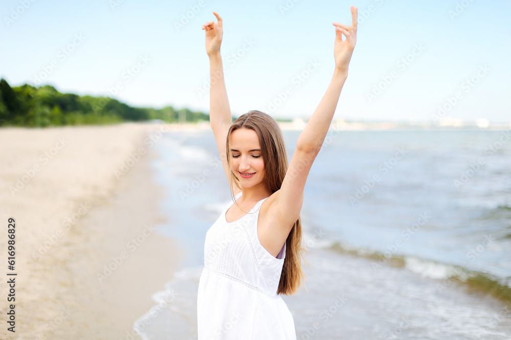 Happy smiling woman in free happiness bliss on ocean beach standing with raising hands. Portrait of a multicultural female model in white summer dress enjoying nature during travel holidays vacation Happy smiling woman in free happiness bliss on ocean beach standing with raising hands. Portrait of a multicultural female model in white summer dress enjoying nature during travel holidays vacation