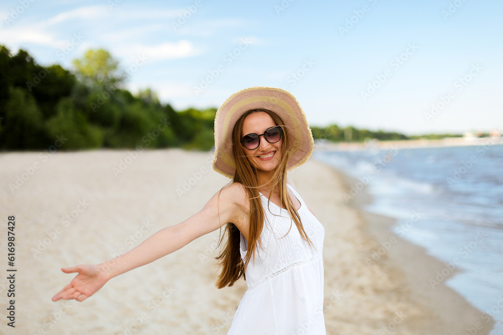 Happy smiling woman in free happiness bliss on ocean beach standing with a hat, sunglasses, and open hands. Portrait of a multicultural female model in white summer dress enjoying nature during travel