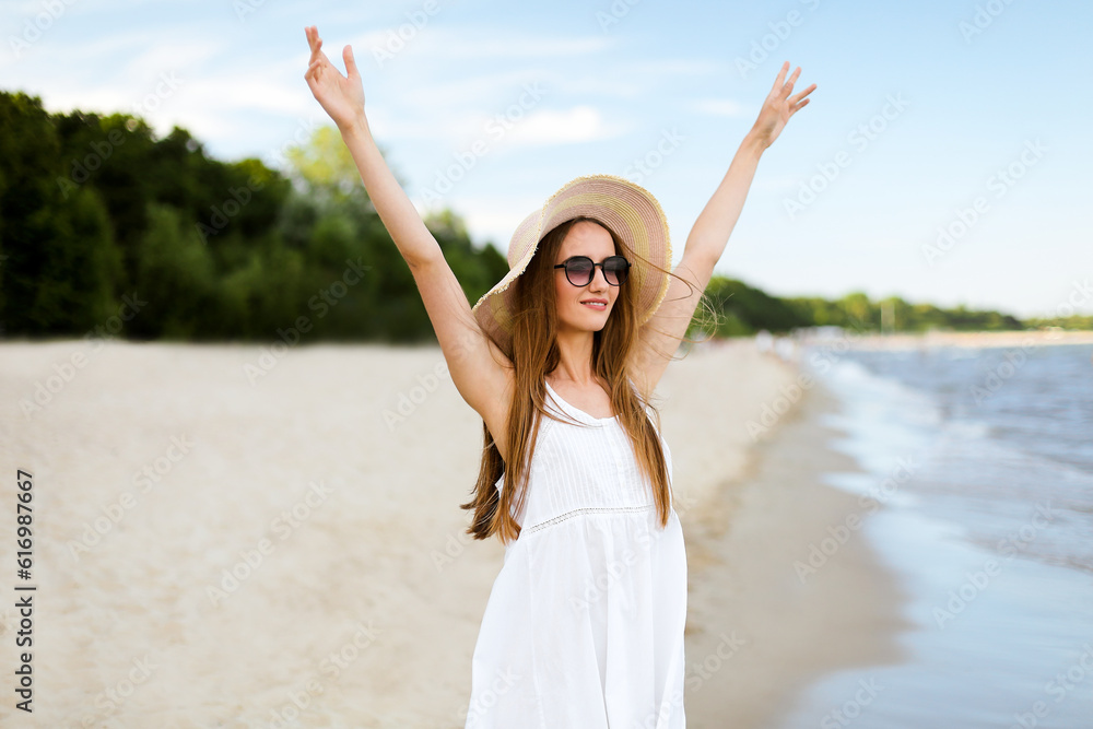 Happy smiling woman in free happiness bliss on ocean beach standing with a hat, sunglasses, and rasing hands. Portrait of a multicultural female model in white summer dress enjoying nature during Happy smiling woman in free happiness bliss on ocean beach standing with a hat, sunglasses, and rasing hands. Portrait of a multicultural female model in white summer dress enjoying nature during