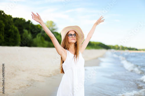 Happy smiling woman in free happiness bliss on ocean beach standing with a hat, sunglasses, and rasing hands. Portrait of a multicultural female model in white summer dress enjoying nature during