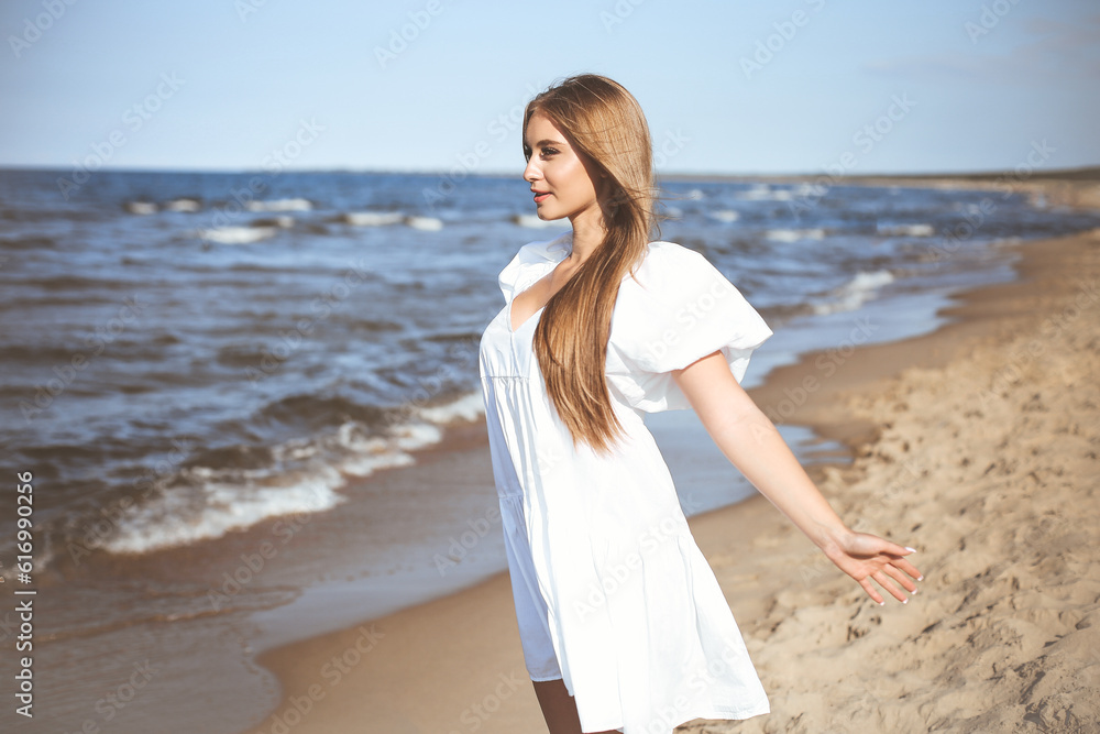 Happy smiling beautiful woman is on the ocean beach in a white summer dress, open arms