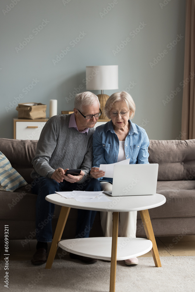 Serious old family sit in living room counting family budget use laptop and calculator, pay bills through e-bank website, review expenses, checks monthly payments. Finances management of older couple Serious old family sit in living room counting family budget use laptop and calculator, pay bills through e-bank website, review expenses, checks monthly payments. Finances management of older couple