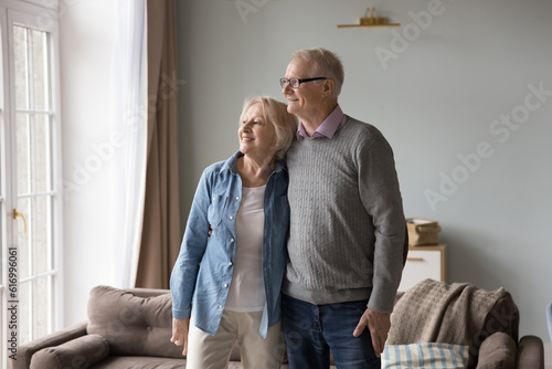 Smiling aged thoughtful wife and husband hugging look out window in living room dreaming about future relish memories standing at new own house. Bank loan for seniors, investment in property, holiday