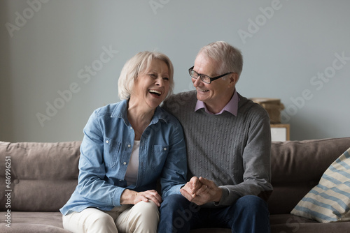 Loving senior man in glasses holds hands his beloved laughing wife, enjoy talk and time together sit on couch, joke, tell jokes, giggling spend pastime at home, enjoy harmony and happy retired life