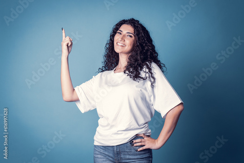 Shot of happy confident curly woman with toothy smile, wears casual basic solid white t-shirt, expresses good emotions, enjoys nice day, isolated over blue background. Expressions