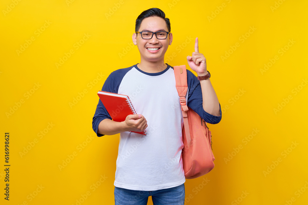 Smart young Asian man student in casual clothes backpack glasses holding book, pointing finger up with great new idea isolated on yellow background. Education in high school university college concept Smart young Asian man student in casual clothes backpack glasses holding book, pointing finger up with great new idea isolated on yellow background. Education in high school university college concept