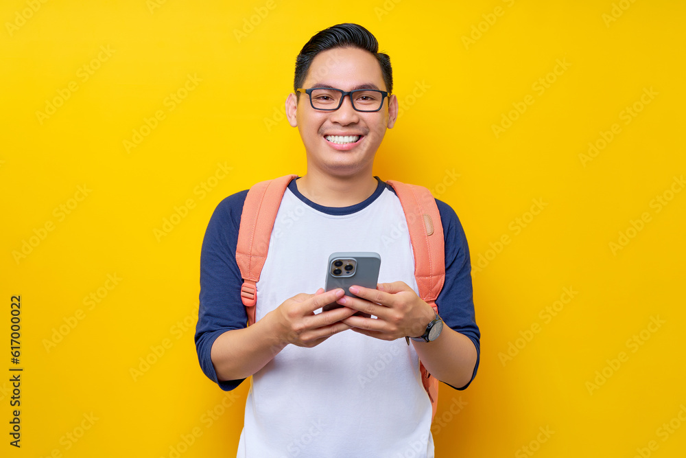 Smiling young Asian man student in casual clothes backpack glasses standing while holding mobile cell phone in hand isolated on yellow background. high school university college concept Smiling young Asian man student in casual clothes backpack glasses standing while holding mobile cell phone in hand isolated on yellow background. high school university college concept