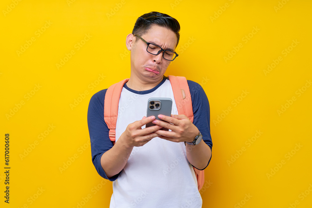 Unhappy young Asian man student in casual clothes backpack glasses standing while using mobile cell phone, chatting online isolated on yellow background. high school university college concept Unhappy young Asian man student in casual clothes backpack glasses standing while using mobile cell phone, chatting online isolated on yellow background. high school university college concept