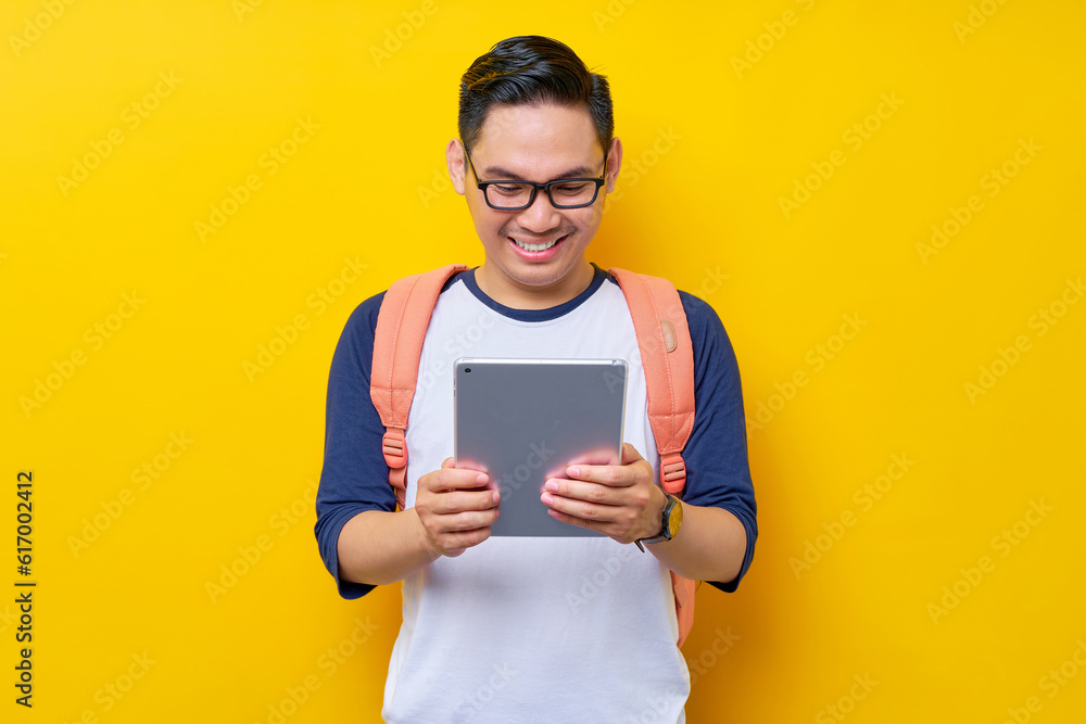 Cheerful young Asian man student in casual clothes glasses backpack using digital tablet computer for studying, reading e-books isolated on yellow background. high school university college concept Cheerful young Asian man student in casual clothes glasses backpack using digital tablet computer for studying, reading e-books isolated on yellow background. high school university college concept