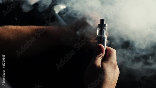 Young vaper man with beard vaping mechanical mod. Guy smokes an electronic cigarette by blowing a smoke vapor. Holds in hand Sitting at the wheel of a car. Space for text