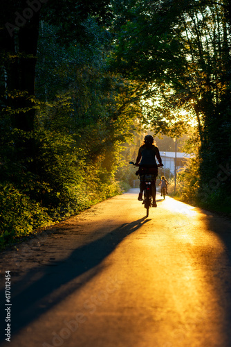 Two cyclists, woman and child, on idyllic bicycle path or bikeway in Hemer Sauerland, Germany. Silhouettes of bikers backlit by low evening summer sun. Golden hour atmosphere on former railway track.