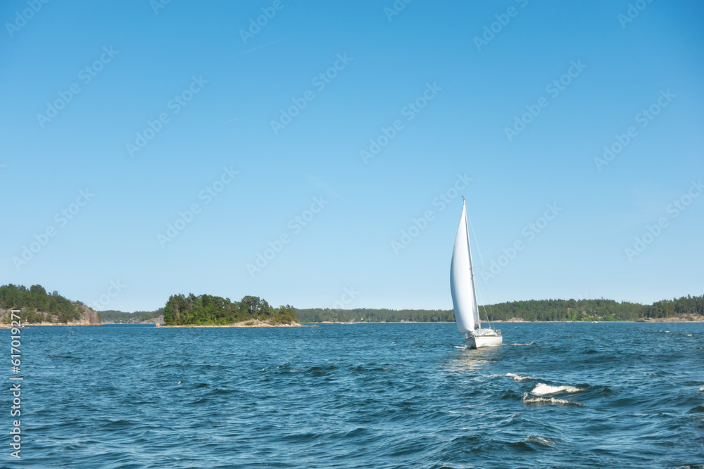 Obraz premium Sailboat with wind in its sails on the blue ocean water in a picturesque archipelago with green islands on a clear summer day for a success and recreational sailing concept