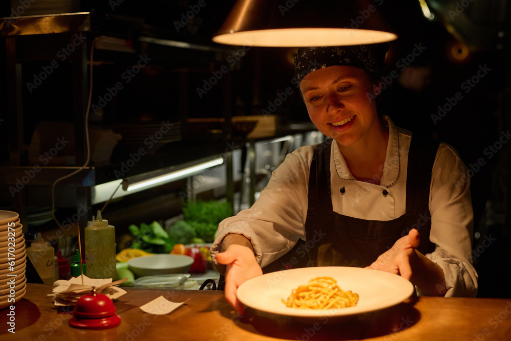 Happy young female worker of cafe in uniform putting plate with appetizing Italian pasta on counter or table while serving it for guests