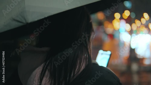Rear view of a girl under an umbrella with a phone walking down at night