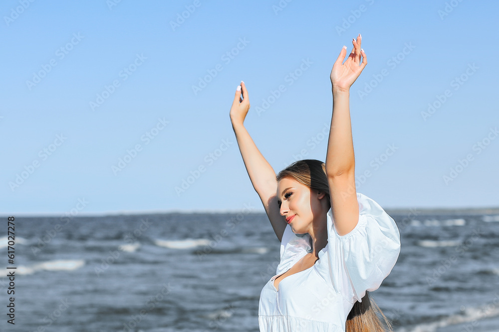 Happy smiling beautiful woman on the ocean beach standing in a white summer dress, raising hands Happy smiling beautiful woman on the ocean beach standing in a white summer dress, raising hands