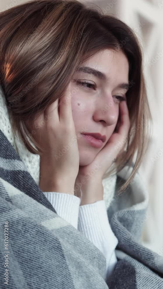 Vertical video. Headache symptom. Feeling flu. Unhealthy woman wrapped in blanket massaging temples suffering pain shivering sitting sofa in light living room at home. Vertical video. Headache symptom. Feeling flu. Unhealthy woman wrapped in blanket massaging temples suffering pain shivering sitting sofa in light living room at home.