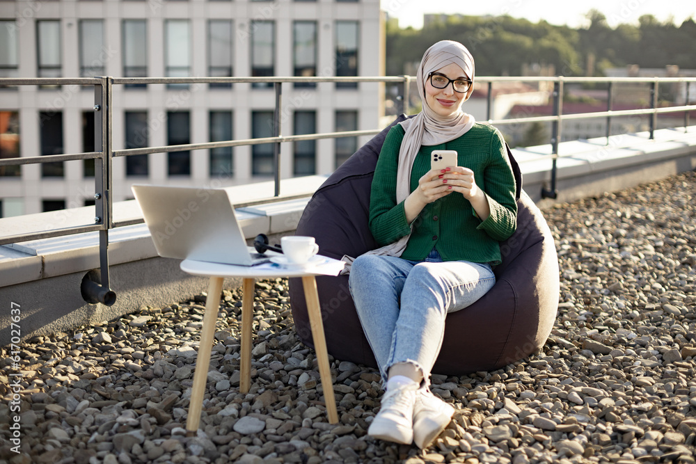 Fototapeta premium Full length view of charming woman in headscarf holding phone while sitting in beanbag near coffee table with gadgets. Delighted muslim person adoring city view in sunlight on panoramic terrace.