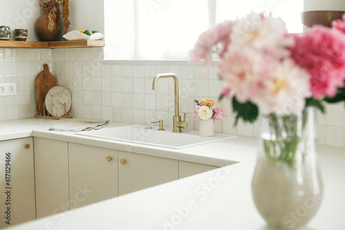Modern kitchen design. Stylish brass faucet and white granite sink, kitchen cabinets with knobs and peonies bouquet in new scandinavian house.  Modern kitchen interior