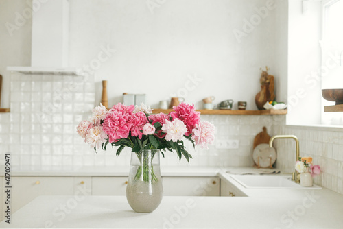 Beautiful peonies in vase on granite countertop on background of stylish white kitchen with wooden shelves and appliances in new house. Modern minimal kitchen interior