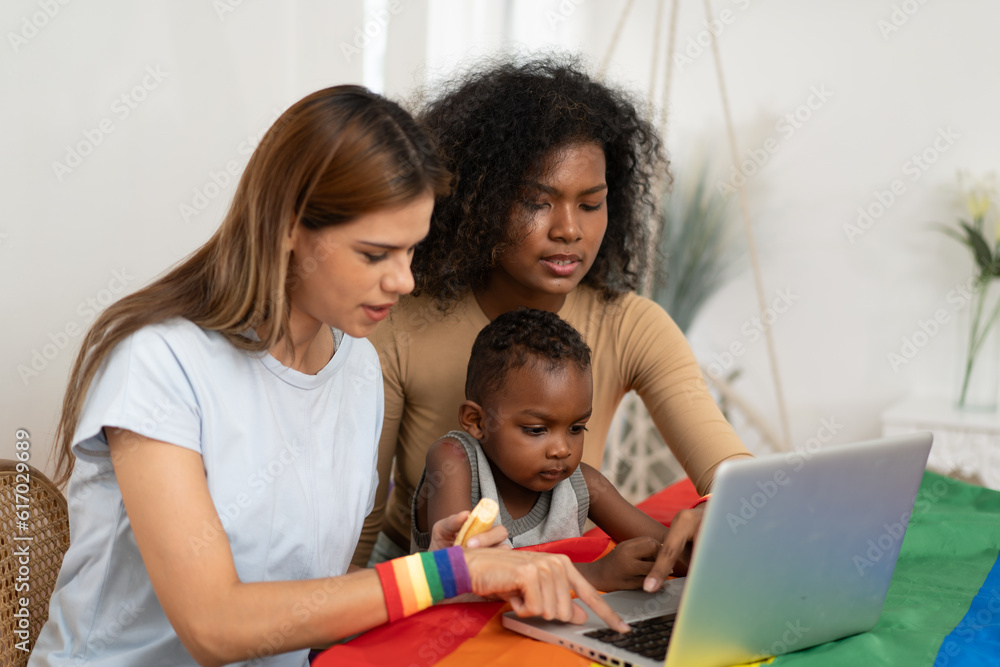 Happy multiethnic women couple having fun with her son watching movie at home. Lesbian mother and child study cyberspace using laptop. Happy female gay family. Homosexual, LGBT relationship lifestyle Happy multiethnic women couple having fun with her son watching movie at home. Lesbian mother and child study cyberspace using laptop. Happy female gay family. Homosexual, LGBT relationship lifestyle