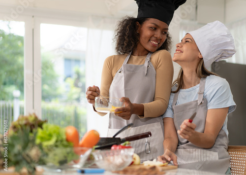 Multiracial lesbian happy cooking in kitchen with girlfriend. Diverse homosexual family preparing food together at home. Cheerful young lgbtq gay woman lover prepare romantic dinner. LGBT lifestyle.