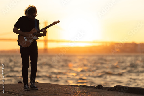 A man stands at the waterfront and playing guitar at bright sunset