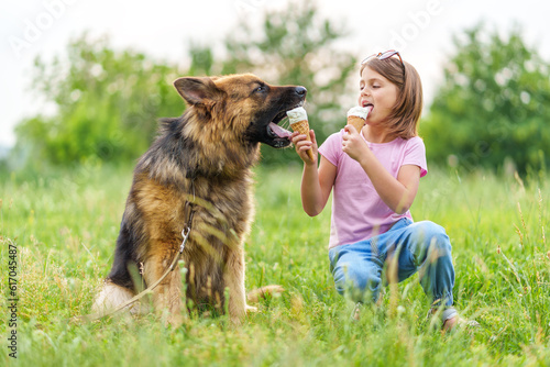 Cute little girl eating ice cream and feeding it to her shepherd dog sitting on the grass in the park in summer. High quality photo, blurred background.