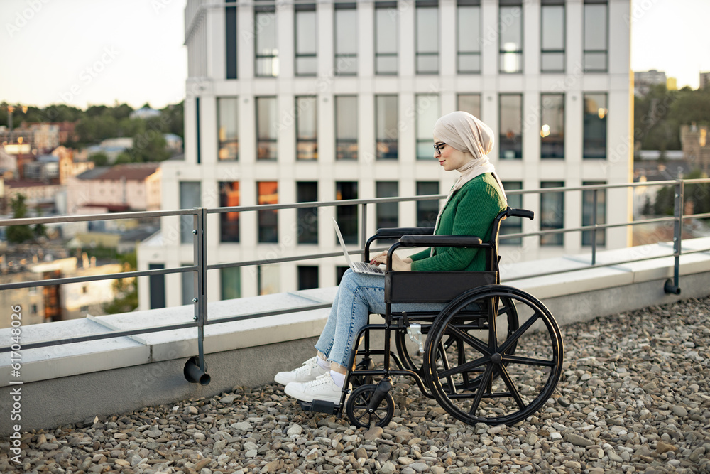 Side view of muslim lady in headscarf using digital device for remote work on panoramic terrace. Well -organized businesswoman in wheelchair searching information over internet application outdoors. Side view of muslim lady in headscarf using digital device for remote work on panoramic terrace. Well -organized businesswoman in wheelchair searching information over internet application outdoors.