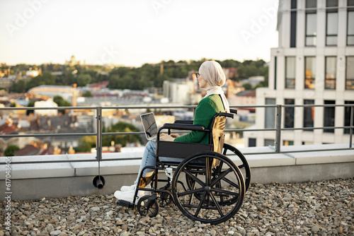 Back view of arabian person in traditional headscarf staring into distance on rooftop against urban background. Relaxed female wheelchair user with laptop admiring picturesque scenery outside. © sofiko14