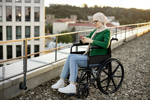 Beautiful person wearing hijab in wheelchair using smartphone while sitting in sunlight on roof terrace. Attractive arabian lady in spectacles writing blog posts on social media sites in fresh air.