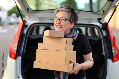 Adult woman with a stack of parcels in her hands with a car with an open trunk in the background.