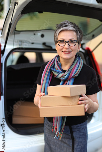 Beautiful adult woman with parcels in her hands with a car with an open trunk in the background.