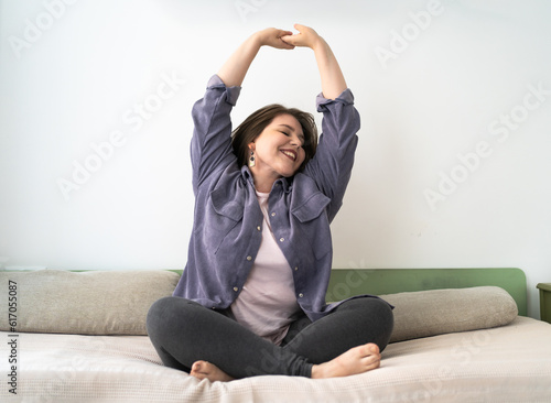 Positive plus size young woman stretching hands up, sitting on sofa with crossed legs. Comfort and relax in weekend.