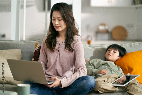 young asian mother sitting on couch working at home while looking after son © imtmphoto