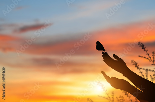 Silhouette hand of woman praying and butterfly flew to the hand with nature on sunrise and orange background with sunlight..