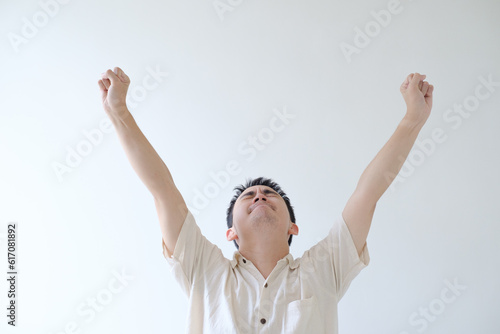 A young Asian man wearing a beige shirt is raising both hands with an excited facial expression. Isolated white background.