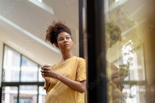 Young African American woman relaxing In modern office looking out of Window with hot drink. © Wasana