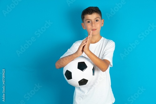 Little hispanic boy wearing white T-shirt holding a football ball clapping and applauding happy and joyful, smiling proud hands together.