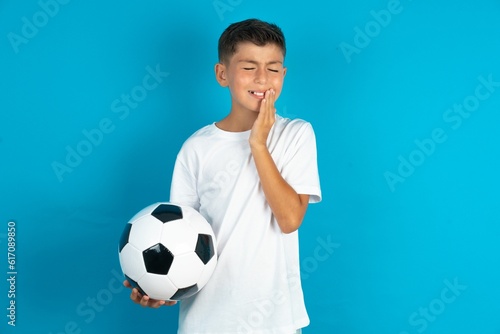 Little hispanic boy wearing white T-shirt holding a football ball touching mouth with hand with painful expression because of toothache or dental illness on teeth. © Jihan