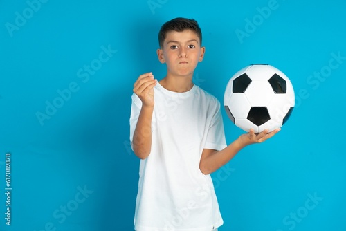 Little hispanic boy wearing white T-shirt holding a football ball doing money gesture with hands  asking for salary payment  millionaire business