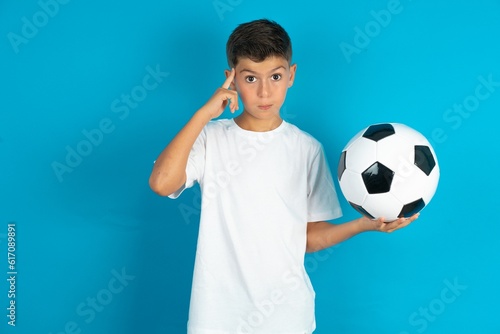 Little hispanic boy wearing white T-shirt holding a football ball confused and annoyed with open palm showing copy space and pointing finger to forehead. Think about it.