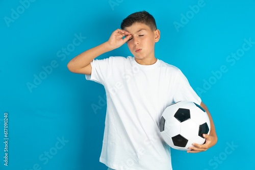 Little hispanic boy wearing white T-shirt holding a football ball having problems, worried and stressed holds hand on forehead.