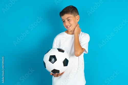 Little hispanic boy wearing white T-shirt holding a football ball Suffering of neck ache injury  touching neck with hand  muscular pain