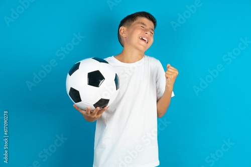Little hispanic boy wearing white T-shirt holding a football ball celebrating surprised and amazed for success with arms raised and eyes closed. Winner concept.
